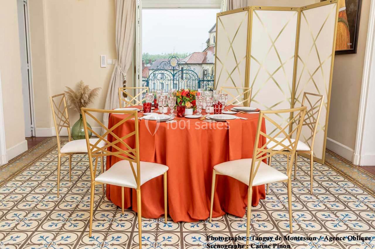 Table ronde dressée avec une nappe orange, entourée de chaises dorées, dans une pièce lumineuse au sol carrelé.