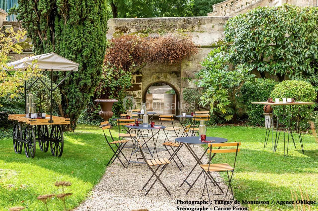 Jardin aménagé avec des tables et chaises en métal et bois, entouré de verdure et d'un mur en pierre avec une arche.
