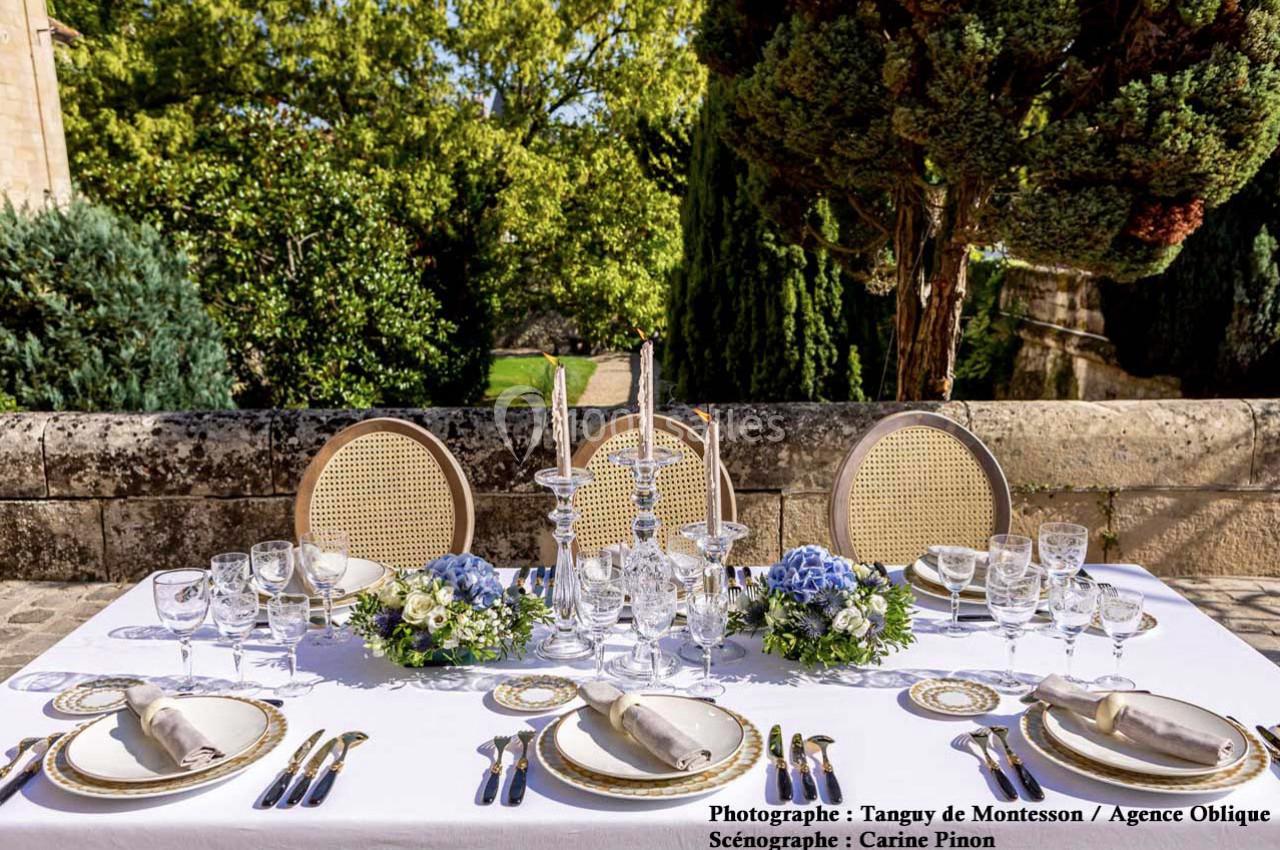 Table élégamment dressée en extérieur avec nappes blanches, vaisselle raffinée et décorations florales bleues et blanches.