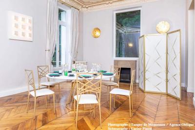 Salle lumineuse avec parquet en bois, table ronde dressée avec nappe rouge et chaises, décorée de moulures au plafond.