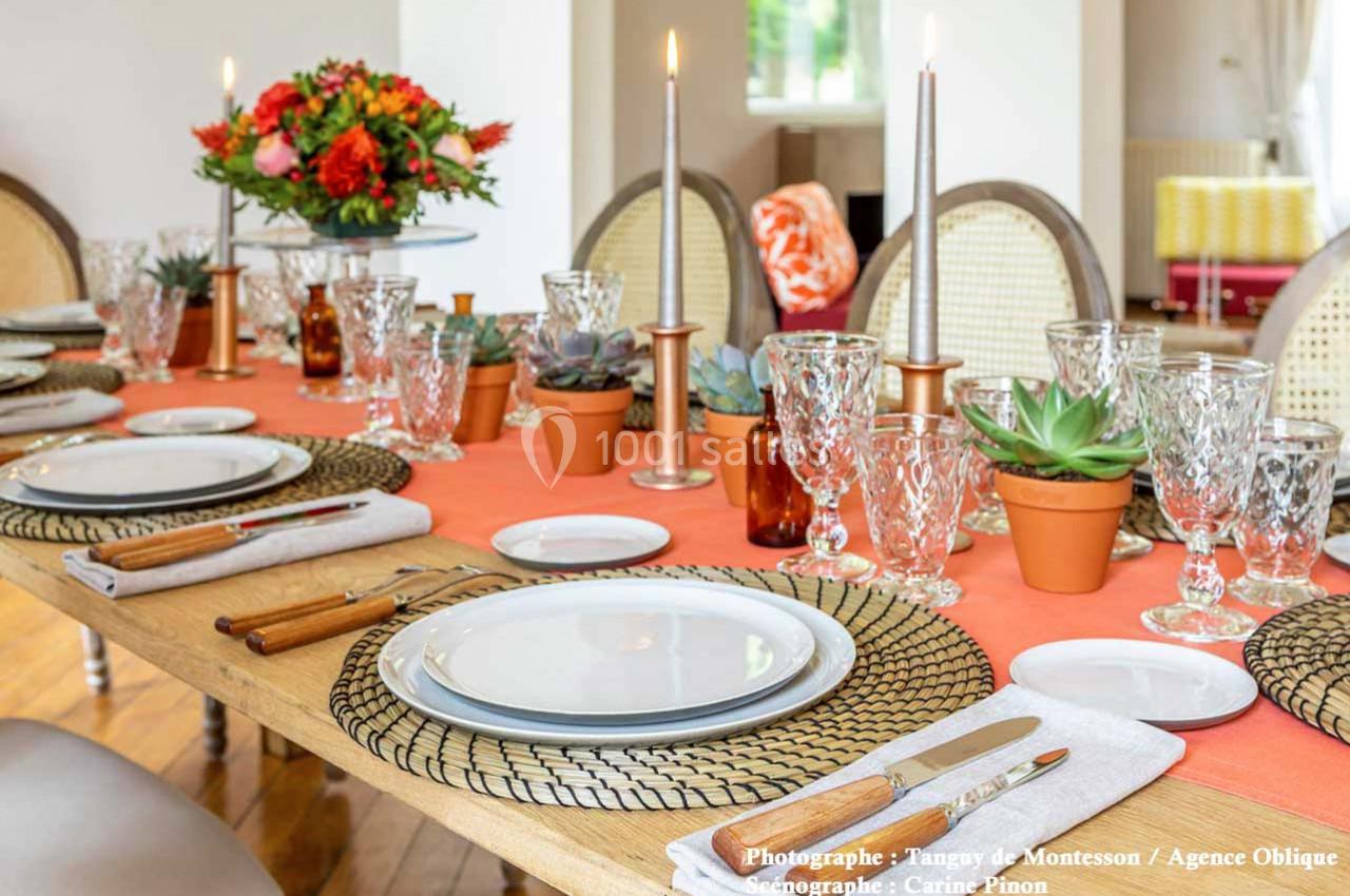 Table dressée avec une nappe orange, vaisselle élégante, bougies allumées et centre de table floral coloré.