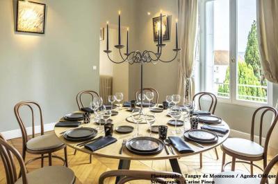 Salle lumineuse avec parquet en bois, table ronde dressée avec nappe rouge et chaises, décorée de moulures au plafond.