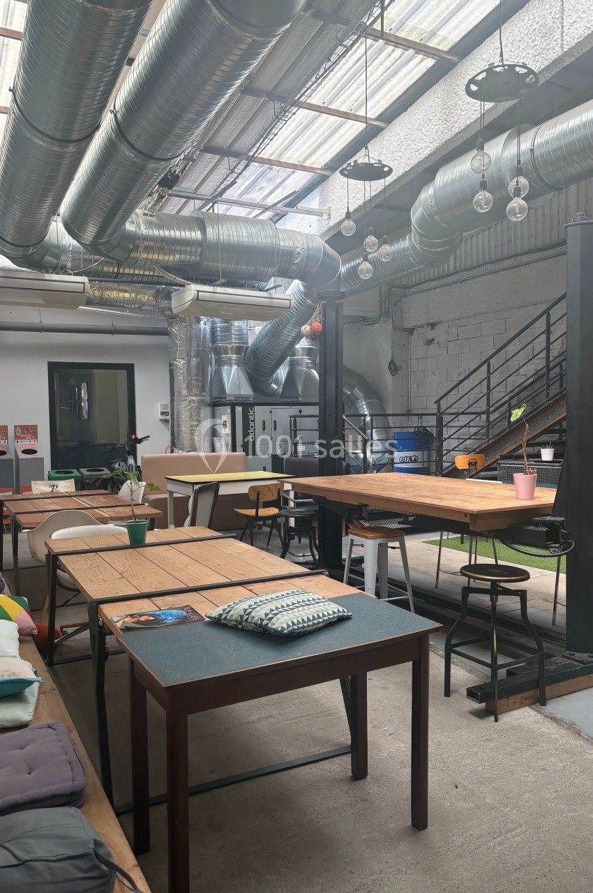 Espace de coworking lumineux avec des tables en bois, des chaises variées et un plafond industriel avec des conduits…