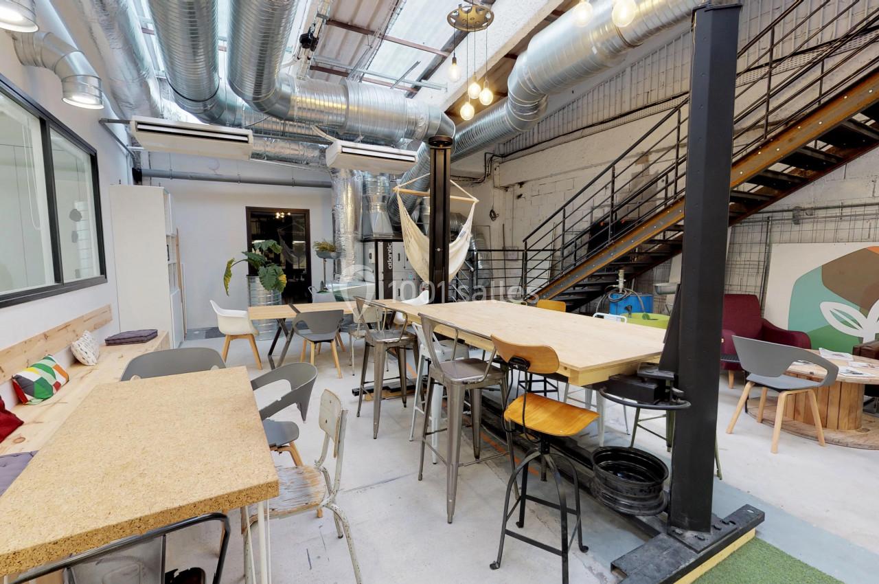 Espace de coworking lumineux avec tables en bois, chaises variées, escalier métallique et décoration industrielle.
