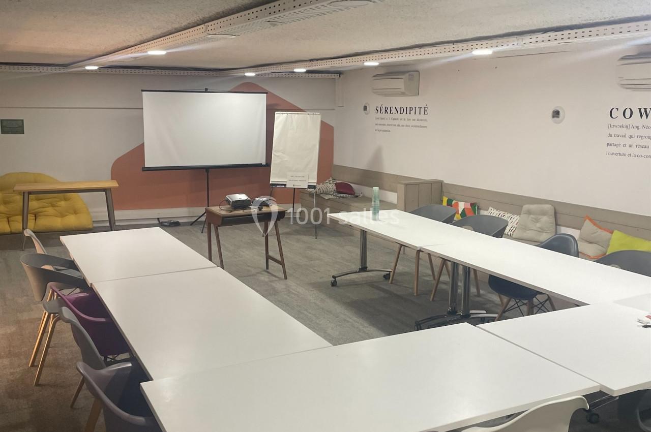 Salle de réunion avec tables disposées en U, chaises colorées, projecteur, écran et décorations murales.