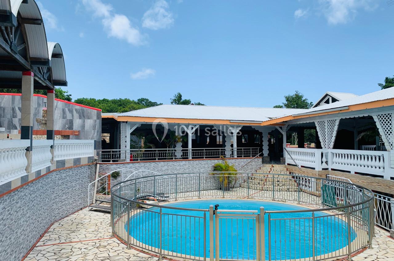 Piscine extérieure entourée de barrières, située dans une cour avec bâtiments aux toits en tôle et structures en bois.