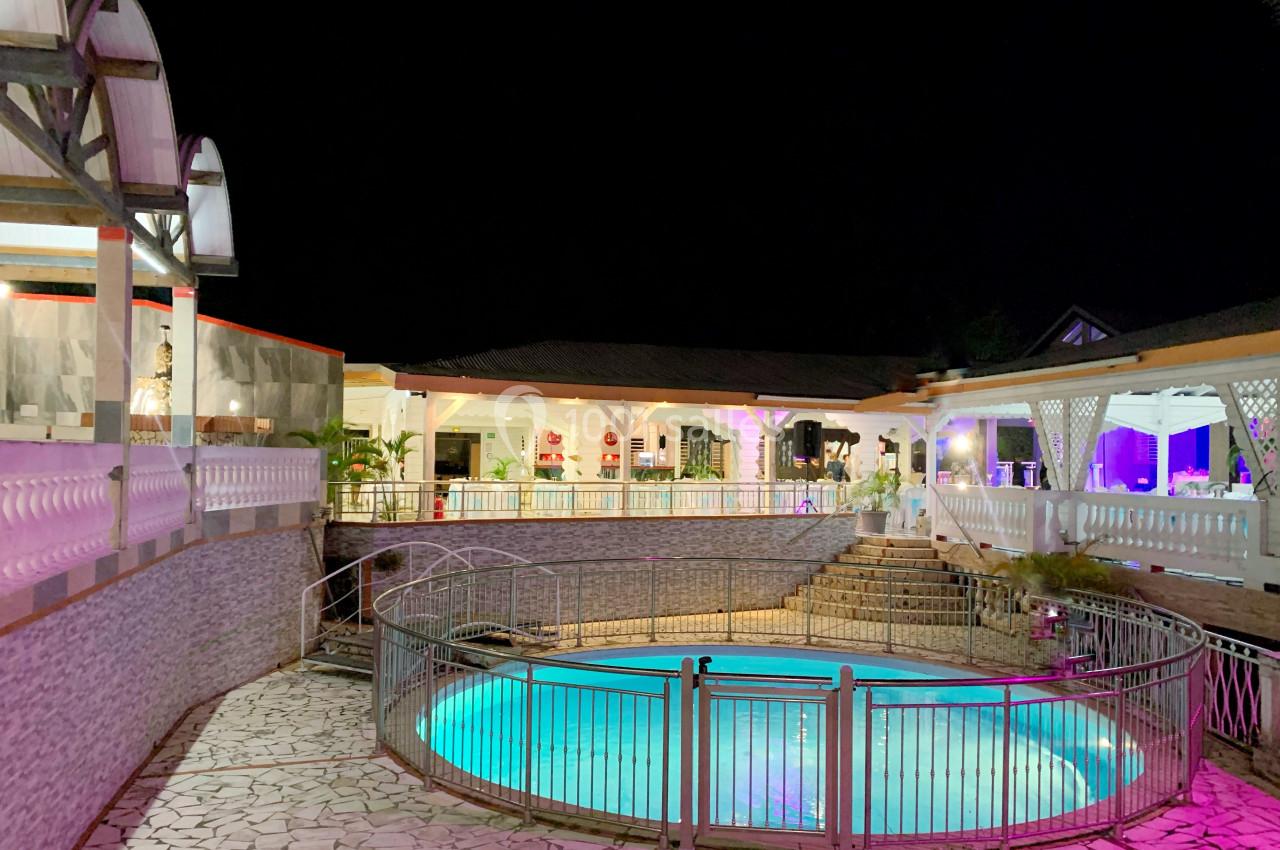 Vue nocturne d'une piscine éclairée entourée de barrières, avec des bâtiments illuminés en arrière-plan.