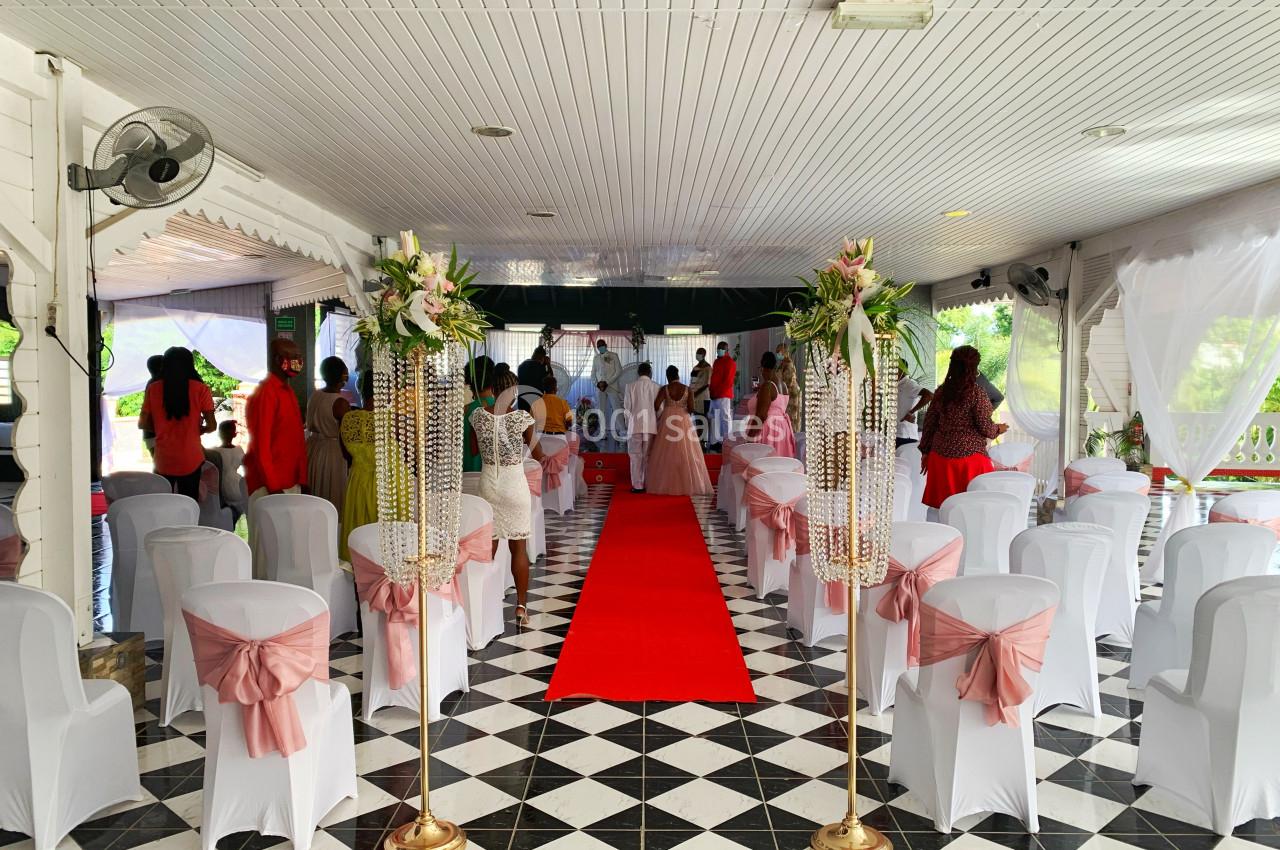 Salle décorée pour une cérémonie avec chaises blanches ornées de rubans roses, tapis rouge et invités debout au fond.