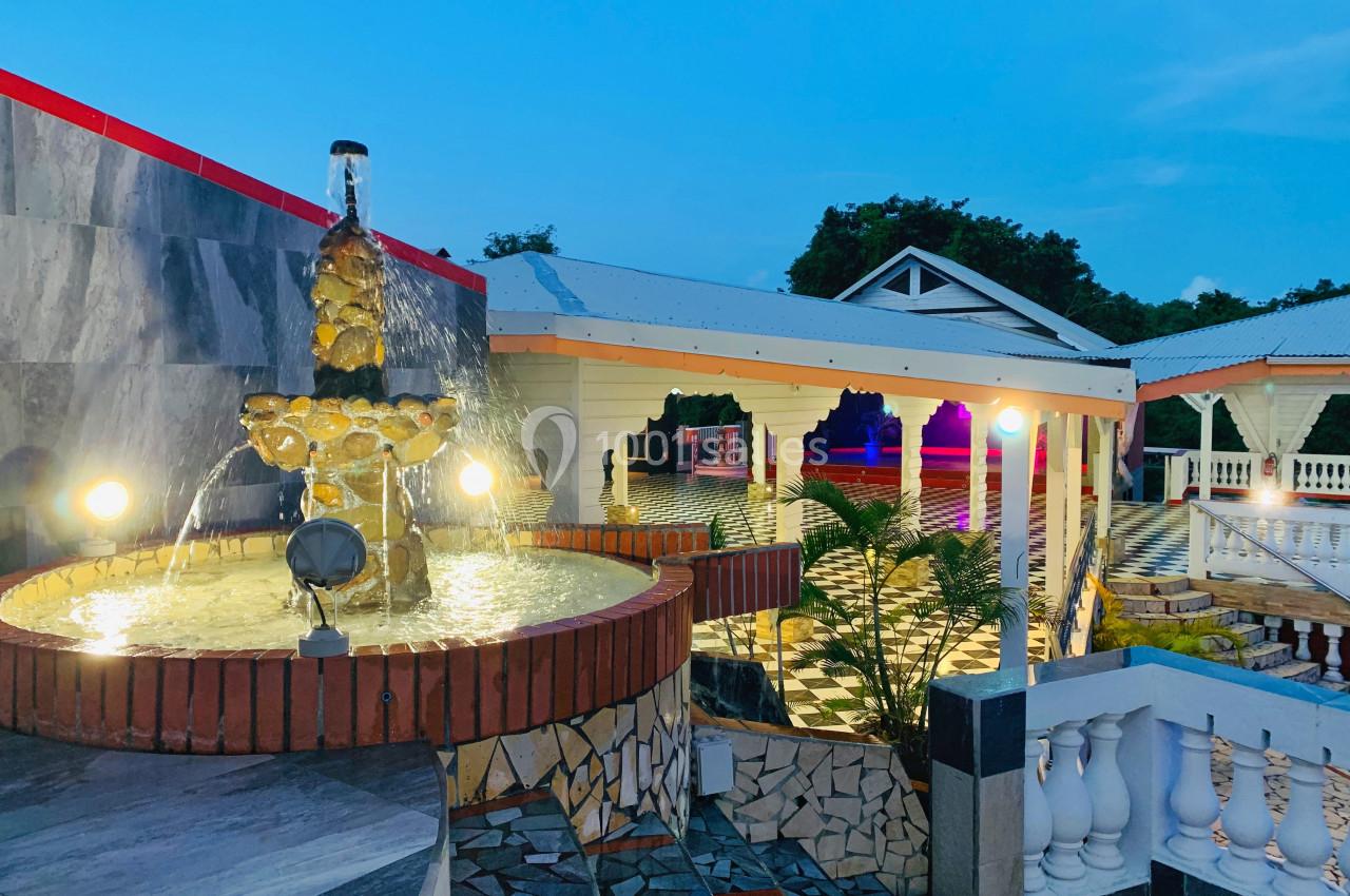 Fontaine illuminée au premier plan avec une terrasse pavée et des bâtiments éclairés en arrière-plan au crépuscule.