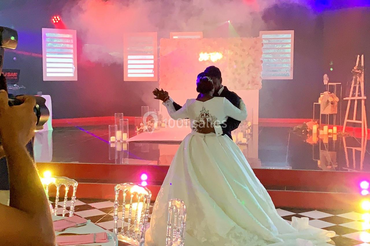 Un couple de mariés danse dans une salle décorée avec des lumières colorées et un sol en damier noir et blanc.