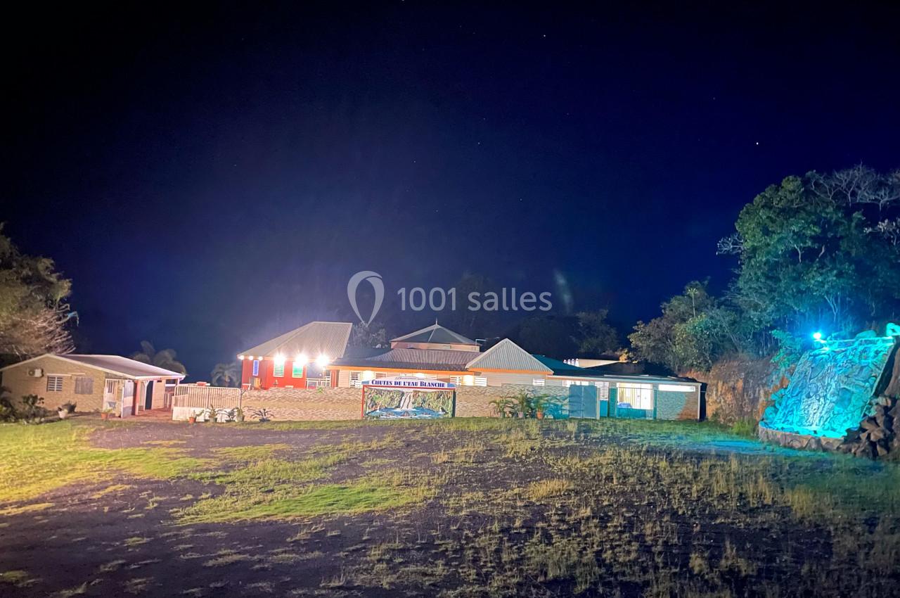 Bâtiments éclairés la nuit avec une cascade illuminée à droite et une pelouse sombre au premier plan.