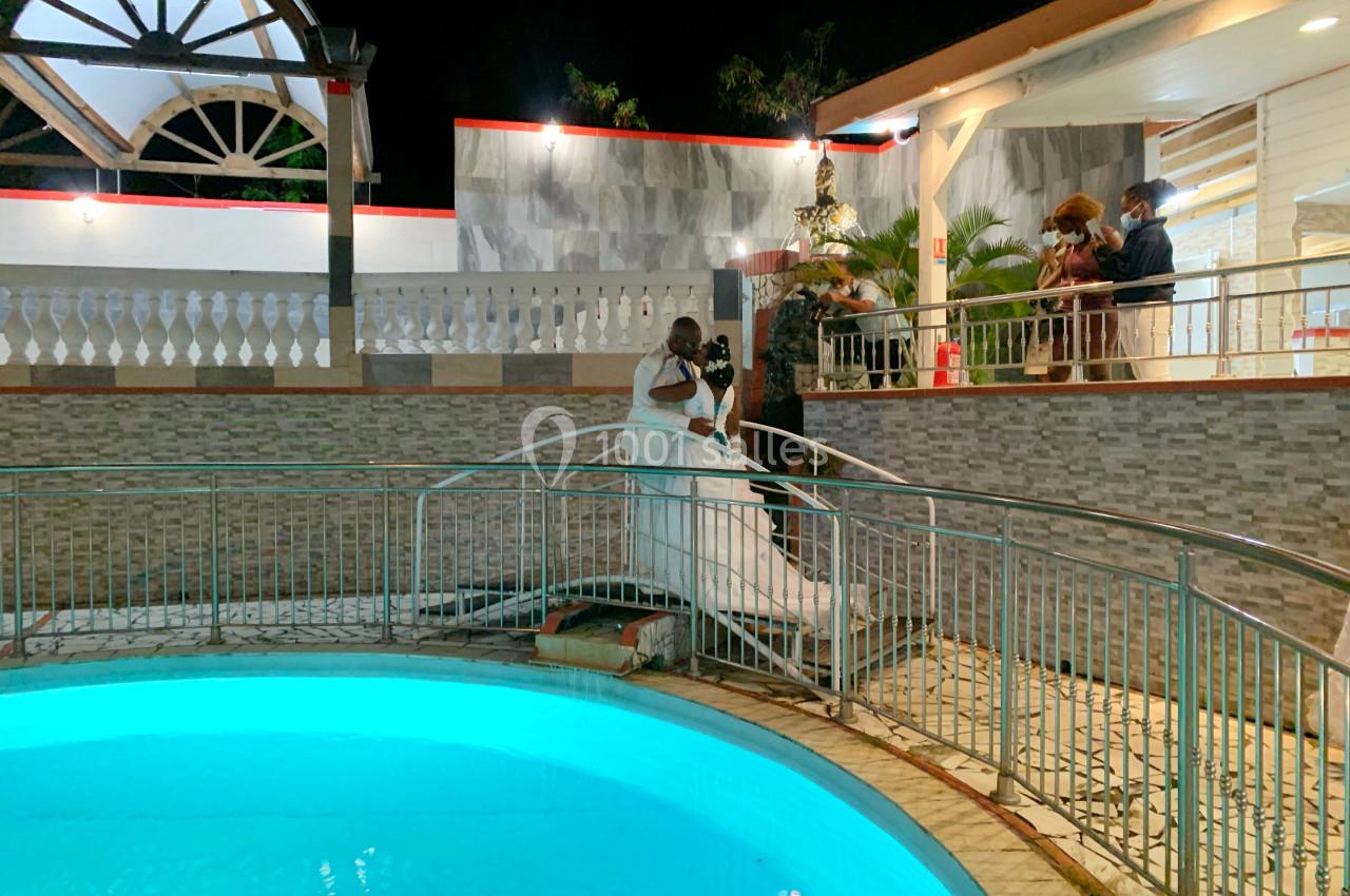 Un couple en tenue de mariage descend un escalier près d'une piscine éclairée, entouré de quelques invités.