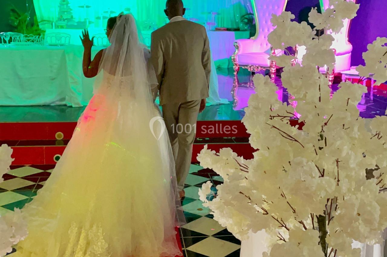 Un couple en tenue de mariage marche sur un tapis rouge dans une salle décorée avec des lumières colorées et des arbres…