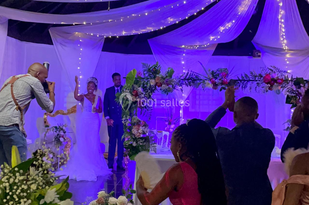 Un couple de mariés pose sur une scène décorée, entouré d'invités prenant des photos dans une salle ornée de fleurs et de…