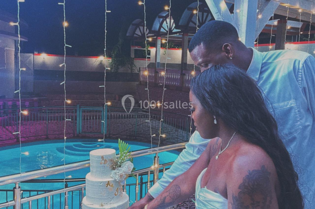 Un couple coupe un gâteau de mariage blanc à plusieurs étages, sous des lumières suspendues près d'une piscine.