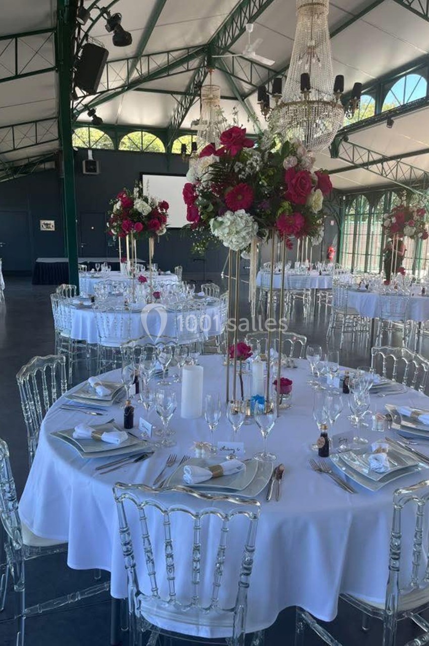 Salle de réception élégante avec tables rondes dressées, décorations florales rouges et blanches, et chandeliers suspendus.