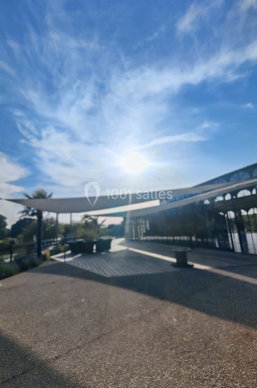 Allée ensoleillée bordée de végétation, avec une structure moderne et un ciel bleu parsemé de nuages.