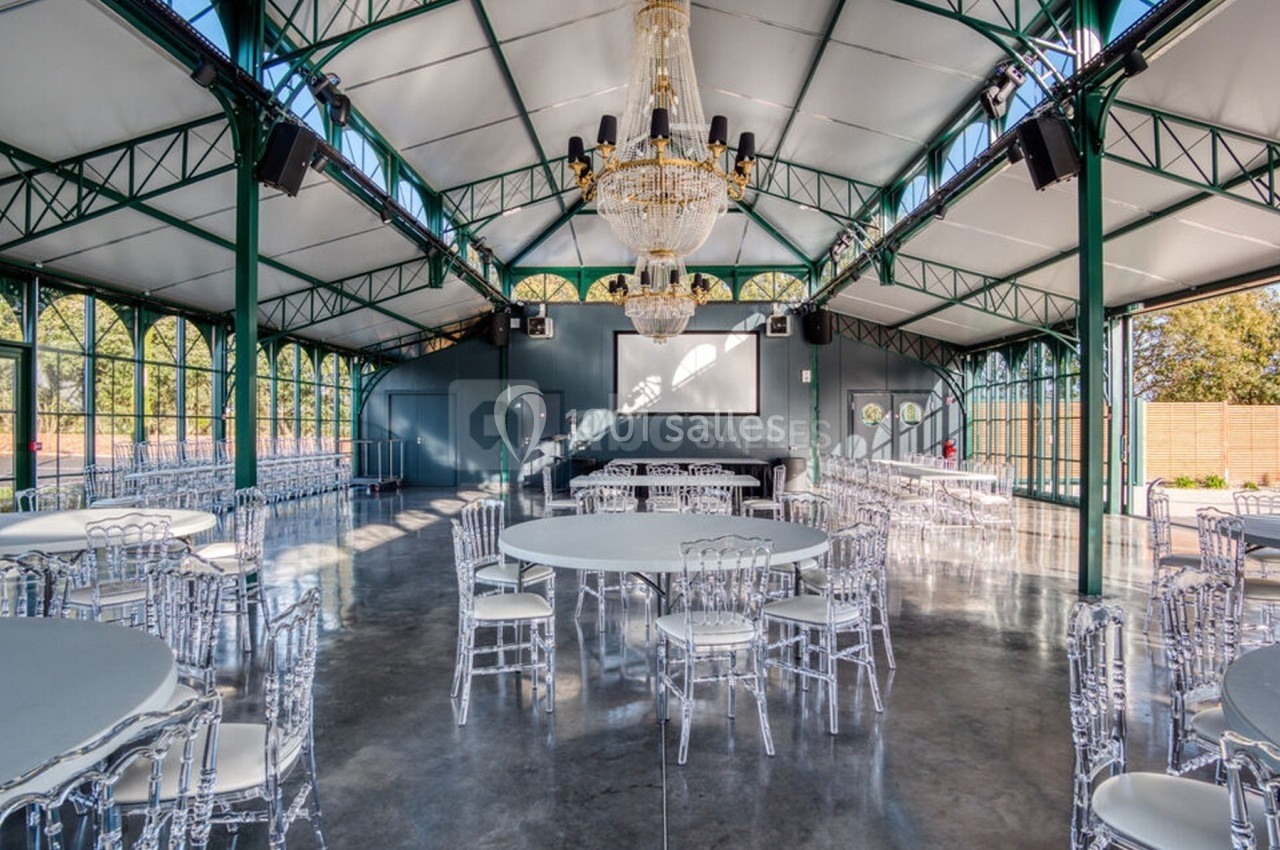 Salle de réception lumineuse avec structure métallique, lustres imposants, tables rondes et chaises transparentes.