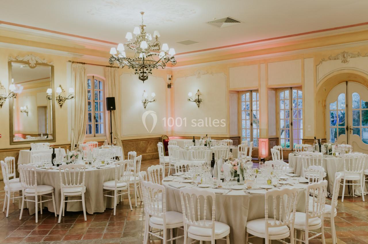 Salle de réception élégante avec tables rondes dressées, chaises blanches et lustres décoratifs.