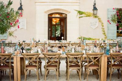 Table en bois décorée de fleurs colorées, verres, assiettes et couverts, installée en extérieur sous une tente.