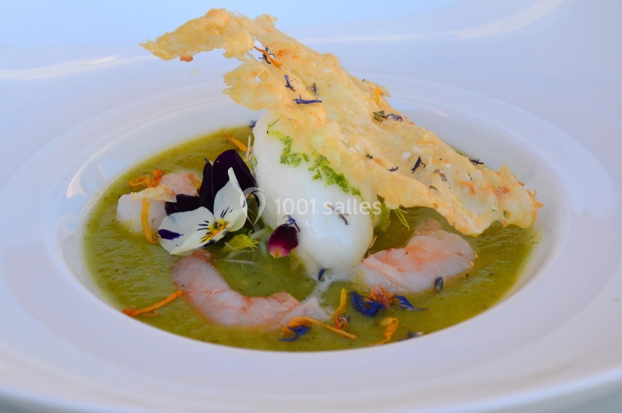 Soupe verte garnie de crevettes, glace, fleurs comestibles et tuile croustillante dans une assiette blanche.