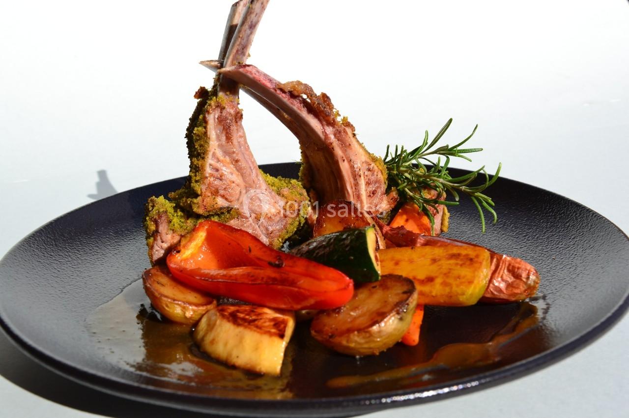Carré d'agneau en croûte d'herbes servi avec légumes rôtis sur une assiette noire.
