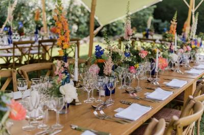 Table en bois décorée de fleurs colorées, verres, assiettes et couverts, installée en extérieur sous une tente.