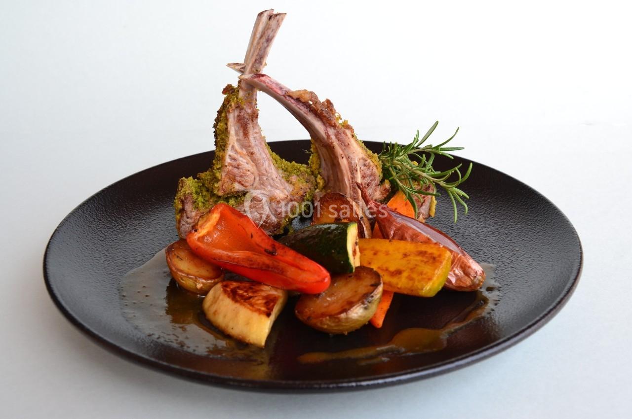 Carré d'agneau en croûte d'herbes servi avec légumes grillés et branche de romarin sur une assiette noire.