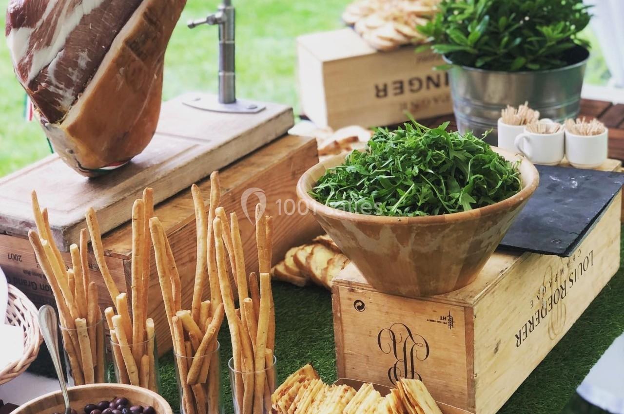 Buffet en plein air avec jambon sur support, grissini, olives, cornichons, roquette et pain sur des caisses en bois.