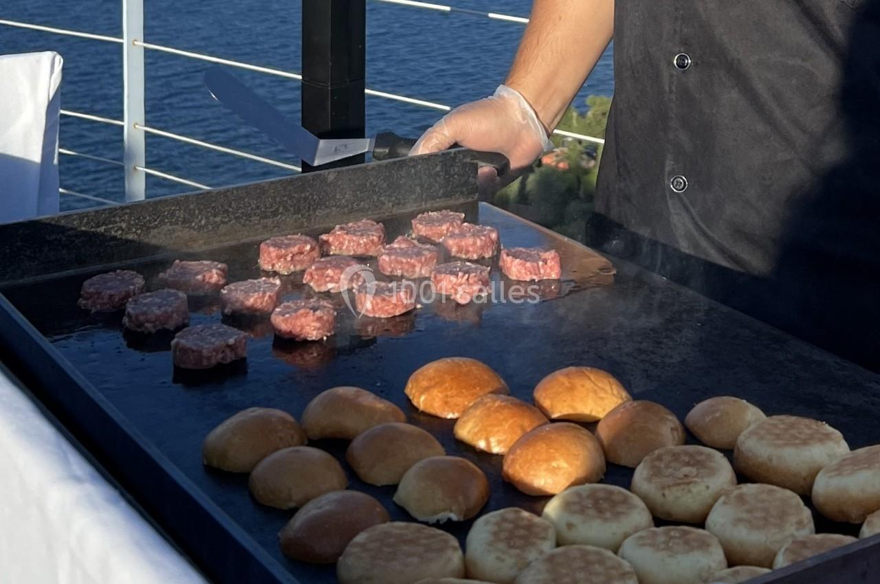 Cuisson de mini-steaks et pains sur une plancha en extérieur, avec vue sur un plan d'eau en arrière-plan.