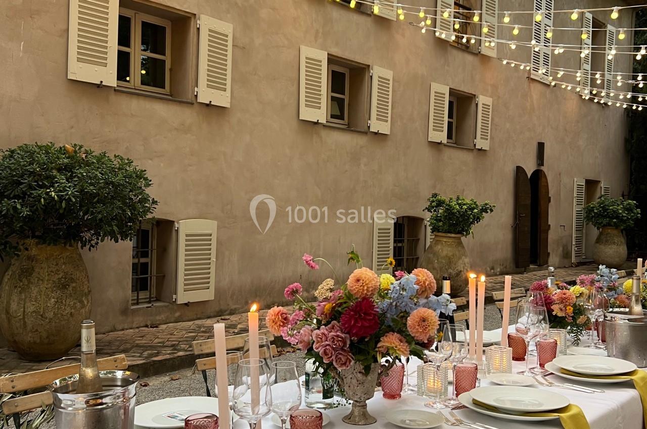 Table dressée pour un dîner en extérieur, décorée de fleurs, bougies et guirlandes lumineuses devant une maison en pierre.