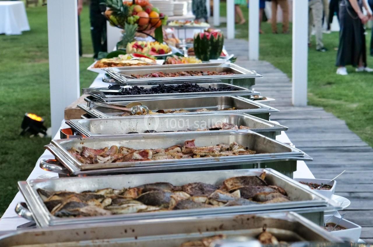 Buffet en plein air avec divers plats chauds et froids, installé sur une table dans un jardin décoré de guirlandes…