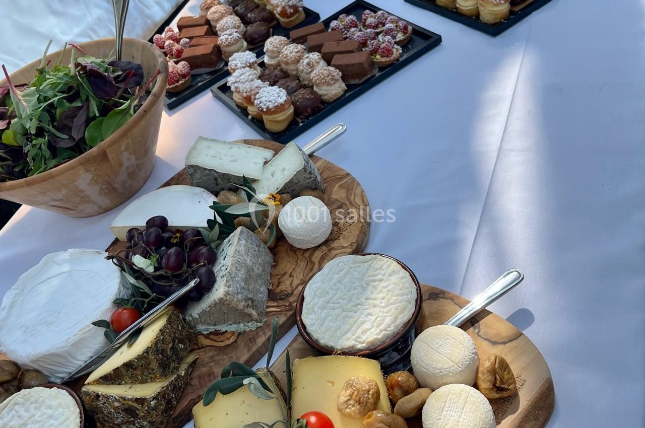 Plateau de fromages variés avec fruits et pains, accompagné de desserts individuels sur une table dressée en extérieur.