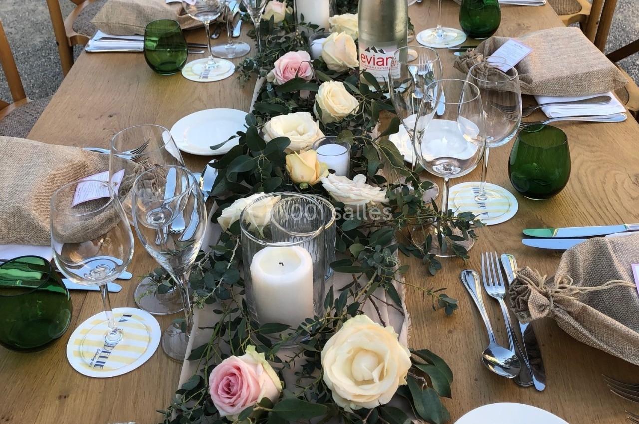 Table en bois décorée pour un repas, ornée de roses, feuillage, bougies blanches et vaisselle disposée avec soin.