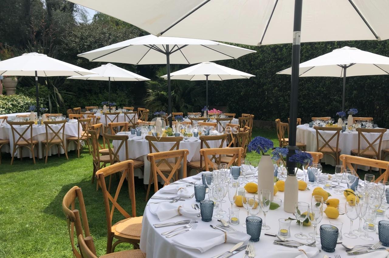 Tables rondes dressées avec nappes blanches, chaises en bois et décorations florales, disposées sous des parasols dans un…