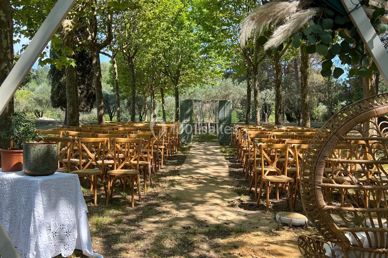 Allée centrale bordée de chaises en bois, décorée de feuillages et de fleurs, menant à une arche dans un cadre naturel.
