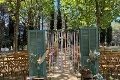 Allée décorée pour une cérémonie en plein air, entourée d'arbres, avec chaises en bois et rubans suspendus.