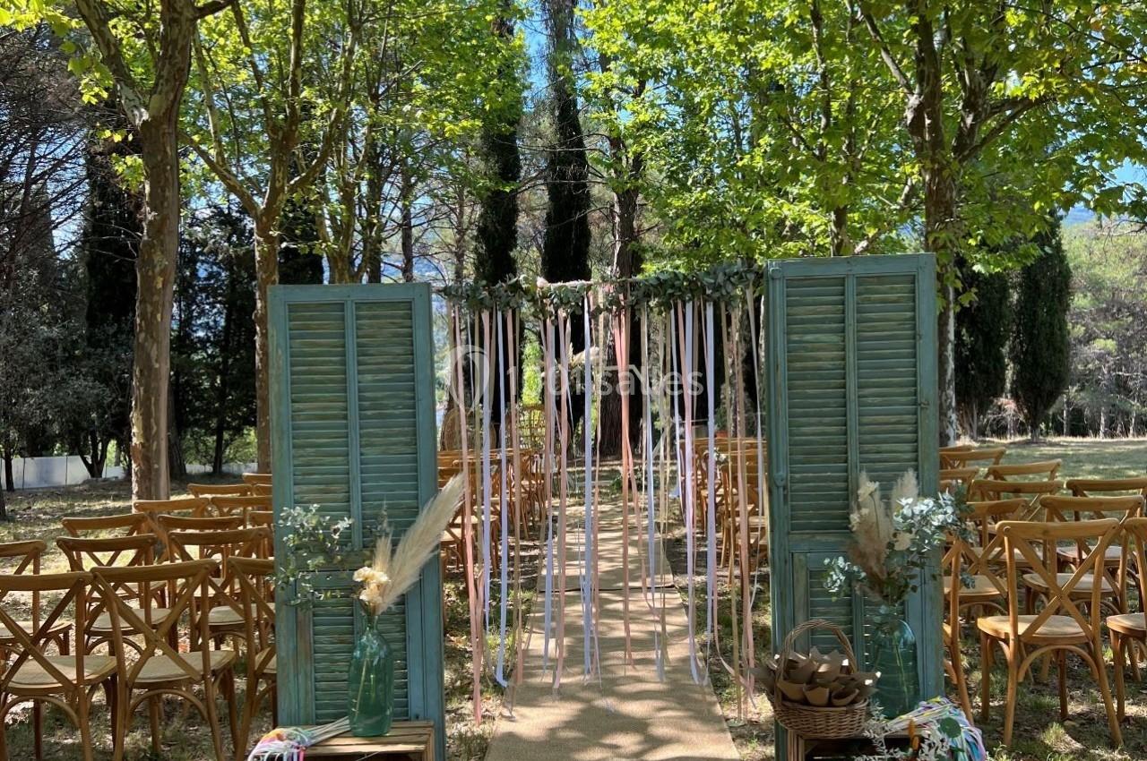 Allée décorée pour une cérémonie en plein air, entourée d'arbres, avec chaises en bois et rubans suspendus.