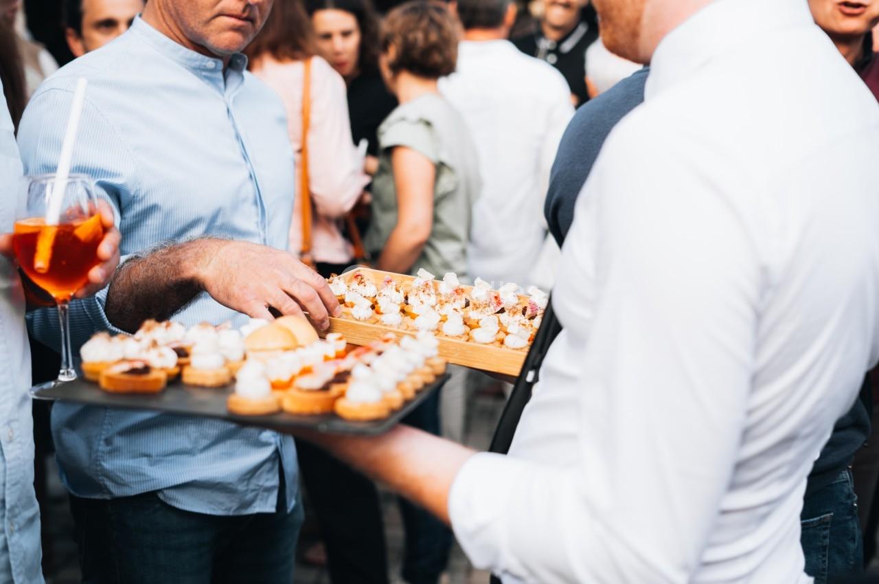 Des personnes debout lors d'un événement, échangeant des plateaux de canapés et de boissons.
