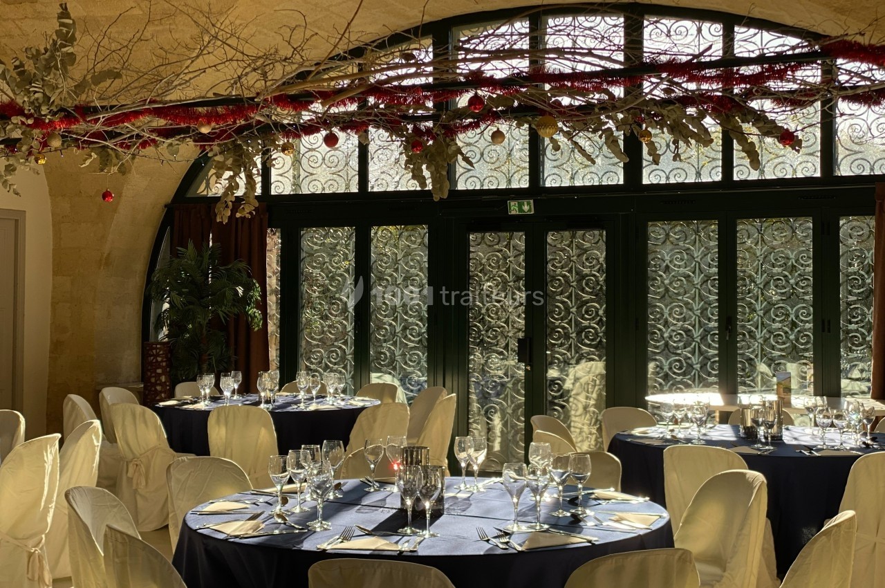 Salle de réception lumineuse avec tables dressées, nappes bleues, chaises blanches et décoration suspendue végétale.