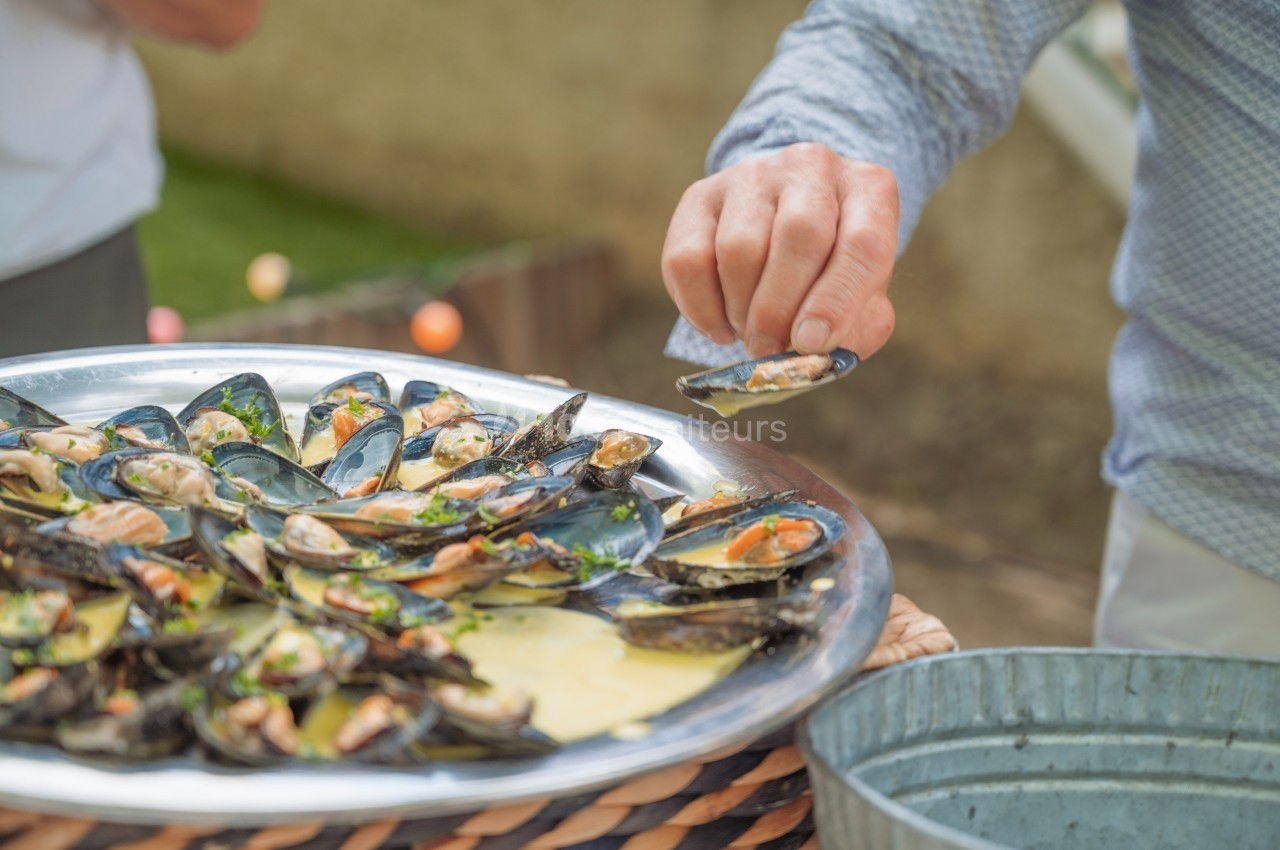 Plateau de moules cuisinées avec sauce, une personne en chemise claire en prend une avec la main.