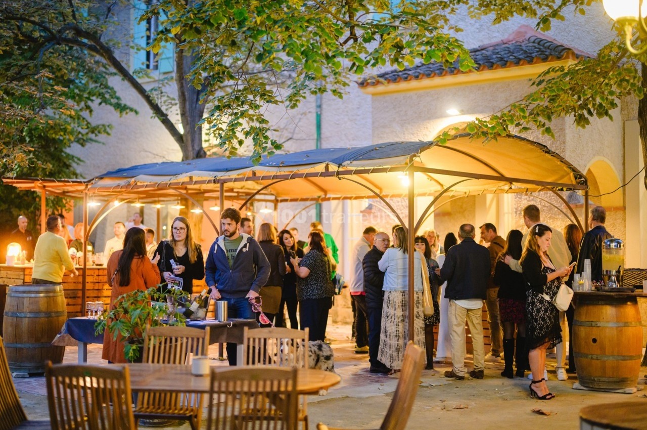 Groupe de personnes discutant sous des tonnelles éclairées dans une cour extérieure, entourée de tables et de végétation.