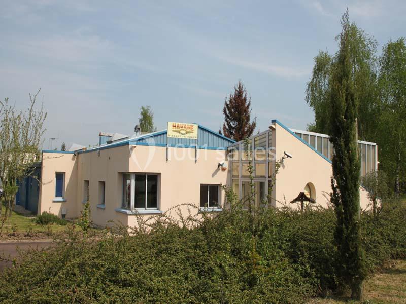 Bâtiment à un étage avec façade beige, toit bleu et végétation environnante sous un ciel dégagé.