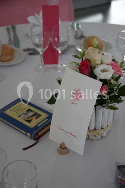 Centre de table avec bouquet de fleurs, menu imprimé, livre et vaisselle disposés sur une nappe blanche.