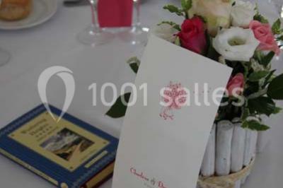 Table dressée avec nappes blanches, couverts, menus et bouquets de fleurs, entourée de chaises en bois dans une salle.