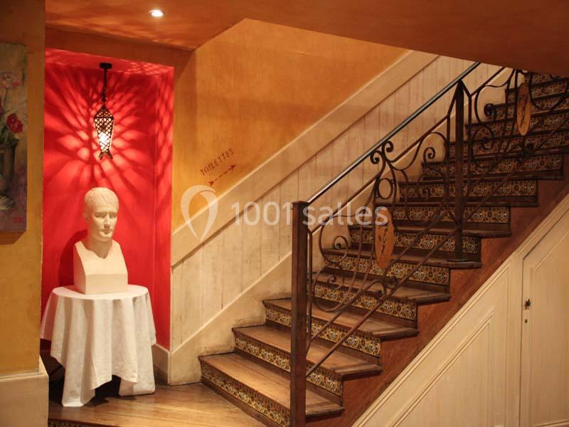 Escalier en bois avec rampe en fer forgé, mur rouge orné d'une applique lumineuse et buste sur une table blanche.