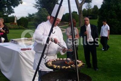 Table dressée avec nappes blanches, couverts, menus et bouquets de fleurs, entourée de chaises en bois dans une salle.