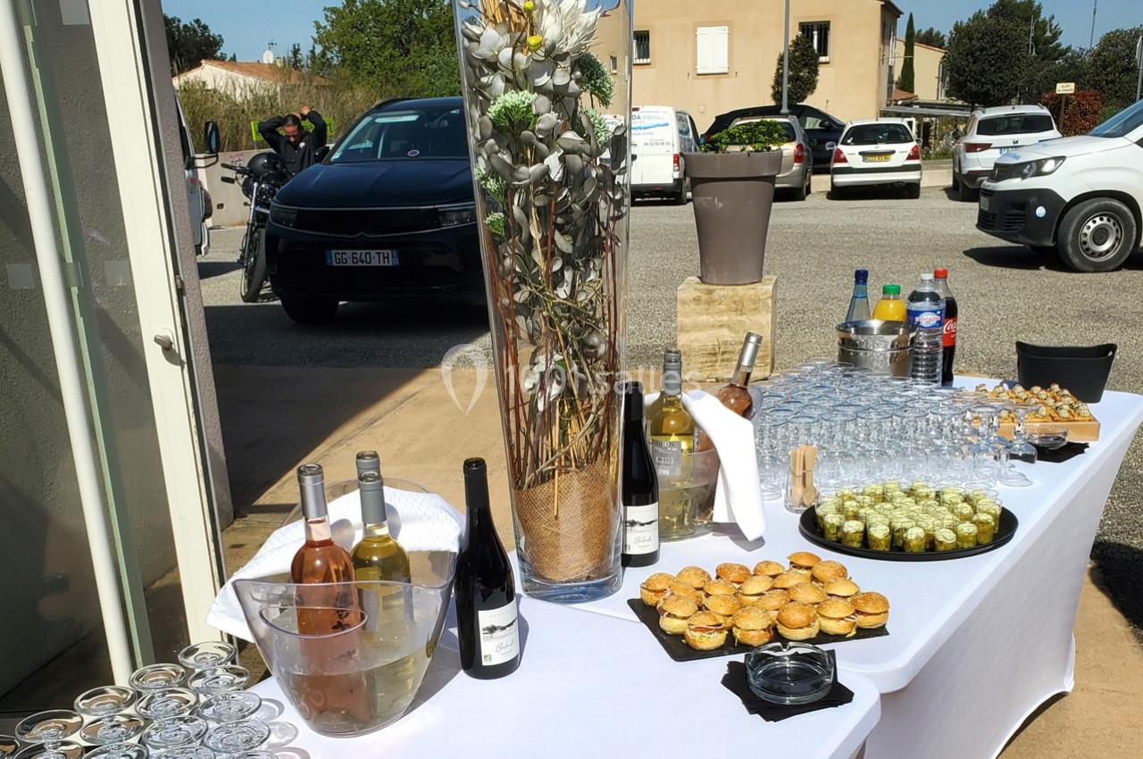Table dressée en extérieur avec boissons, verrines et mignardises, décorée d'un grand vase floral, voitures en arrière-plan.