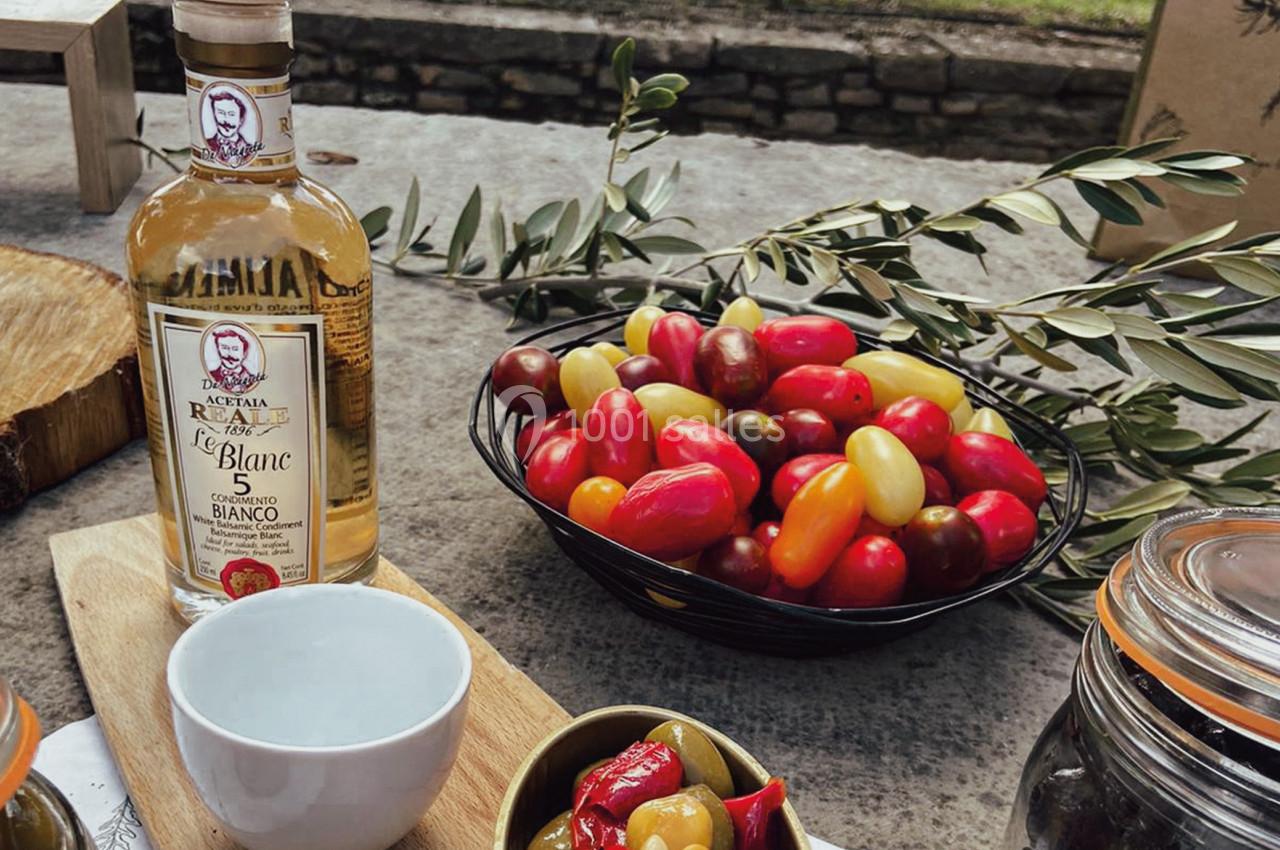 Bouteille de vinaigre blanc, bol de tomates colorées et olives sur une table en extérieur avec des branches d'olivier.