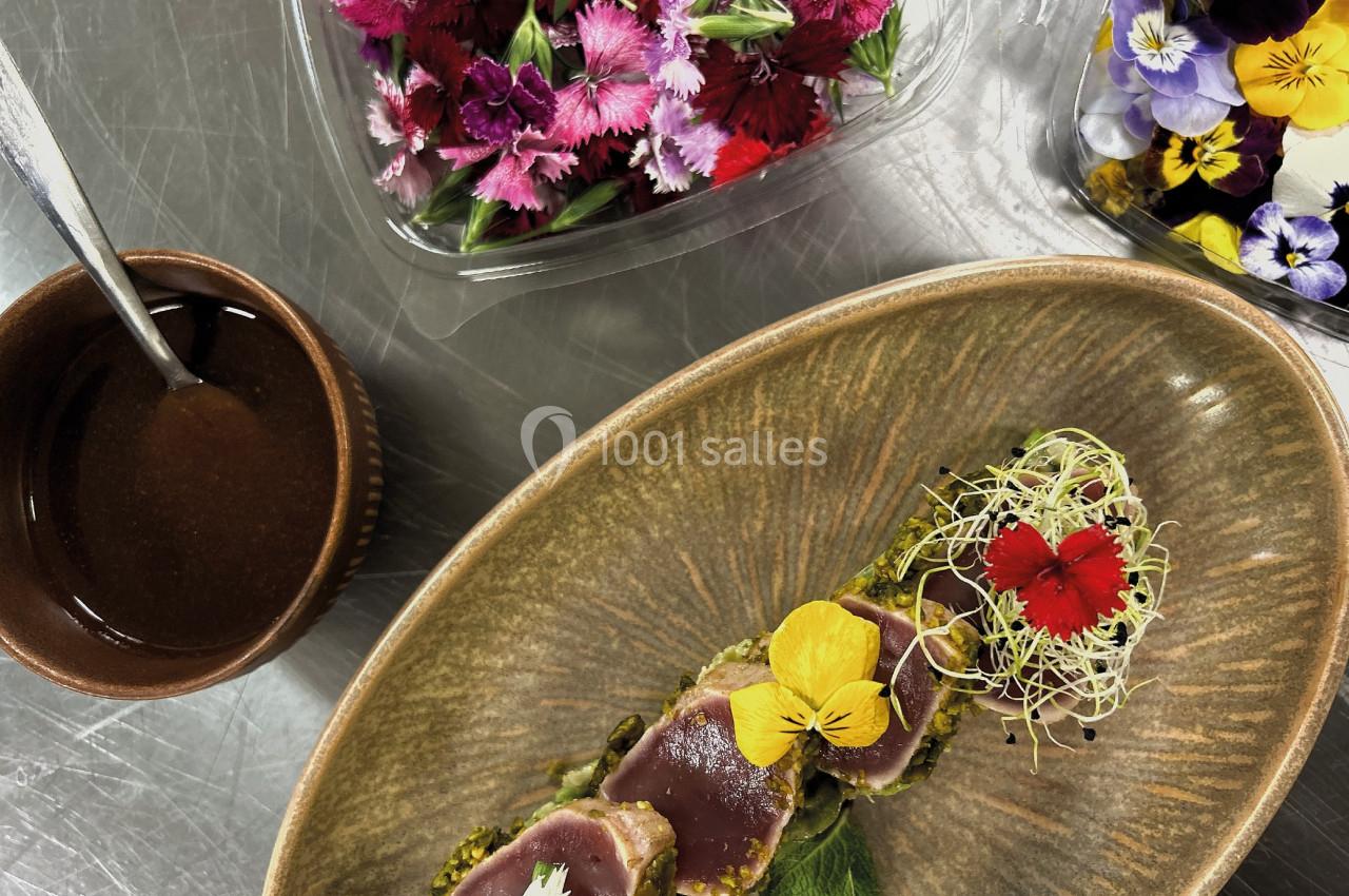 Assiette de thon mi-cuit en croûte, décoré de fleurs comestibles et pousses, avec sauce et fleurs en arrière-plan.